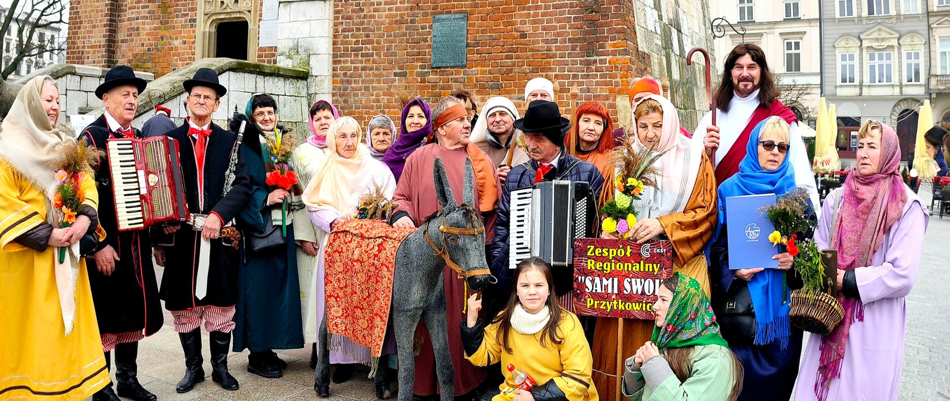 Grupa kilkunastu osób w strojach stylizowanych na biblijne i ludowe stoi przed ceglanym budynkiem na krakowskim rynku. Uczestnicy trzymają palmy i instrumenty, wśród nich widoczny jest mężczyzna przebrany za Jezusa z krzyżem oraz postać siedząca na drewnianym osiołku. Na pierwszym planie znajdują się także dzieci w kolorowych strojach. Część osób trzyma tablicę z napisem „Zespół Regionalny Sami Swoi Przytkowice”.