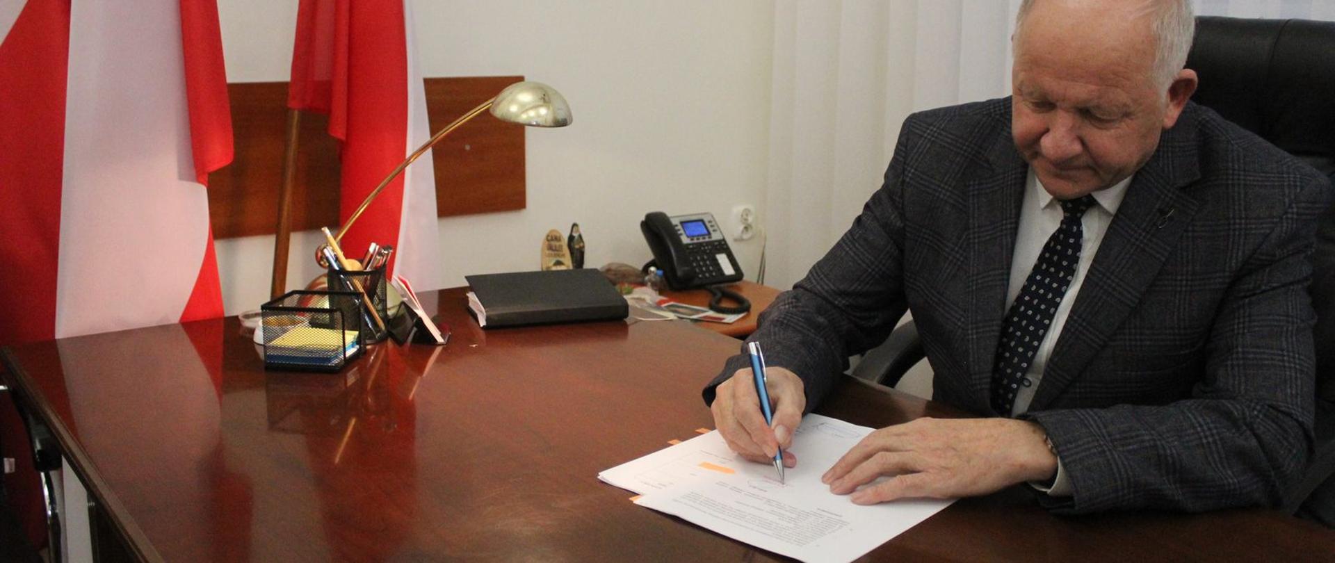 W prawej części fotografii Burmistrz Miasta podpisujący dokumenty, w lewej części flaga Polski biało-czerwona. 