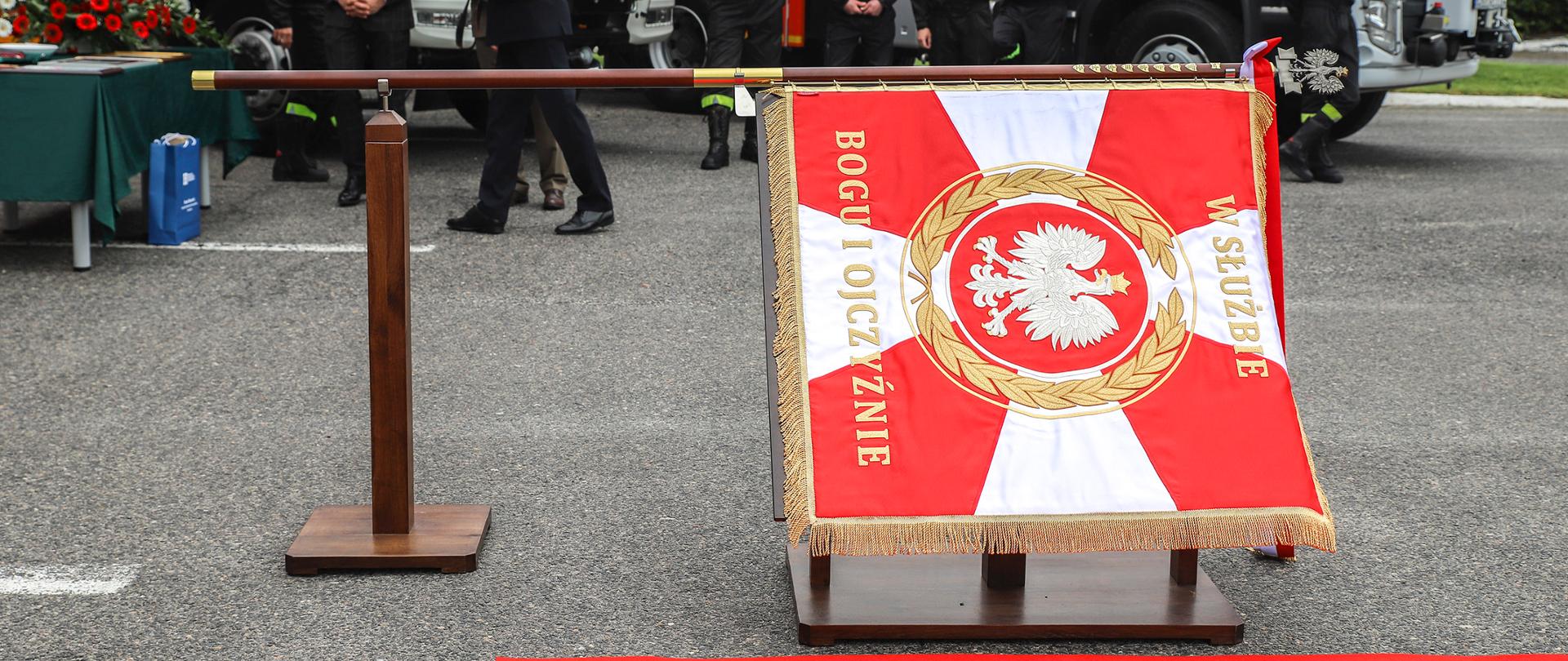 Zdjęcie przedstawia sztandar ułożony poziomo na postumencie. Sztandar w kolorach biało czerwonych został zawieszony na drzewcu ze złotymi okuciami, zwieńczony metalowym orłem w koronie. W centralnej części sztandaru znajduje się godło państwowe otoczone złotym laurem. Nad nim znajduje się napis "w służbie" poniżej godła napis brzmi "Bogu i Ojczyźnie". W tle zdjęcia znajdują się strażacy uczestniczący w wydarzeniu za nimi zaparkowano dwa wozy stażackie.