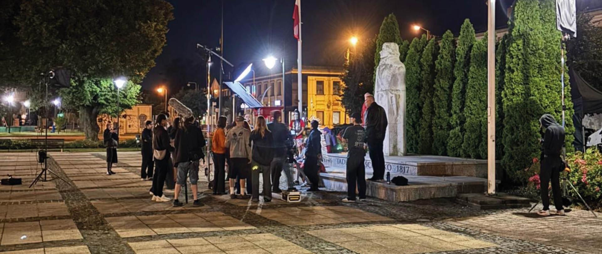 Studenci w nocy na placu Kościuszki przy pomniku, rozstawiony sprzęt filmowy i oświetlenie.