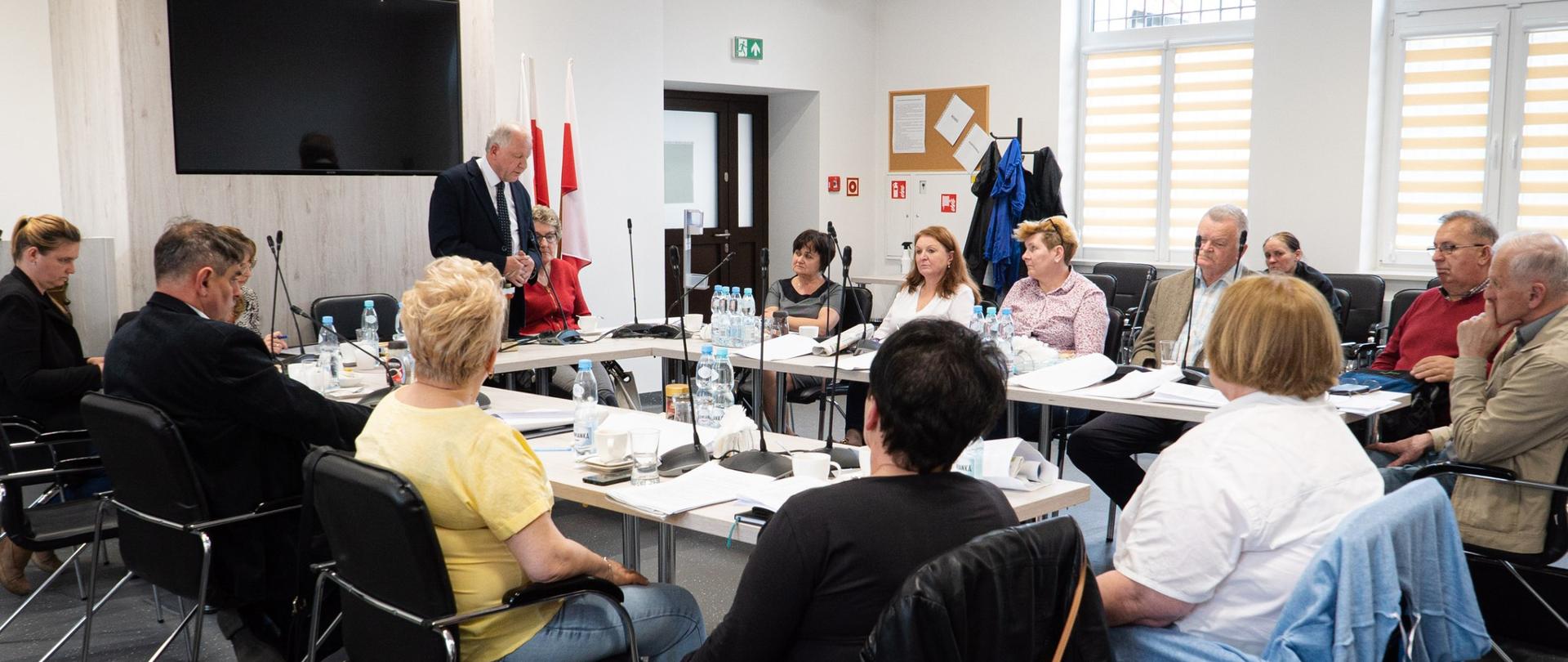 Goście burmistrza siedzą przy stołach ułożonych w podkowę, na których znajdują się mikrofony i dokumenty. Siedzący patrzą na burmistrza, który przemawia na stojąco.