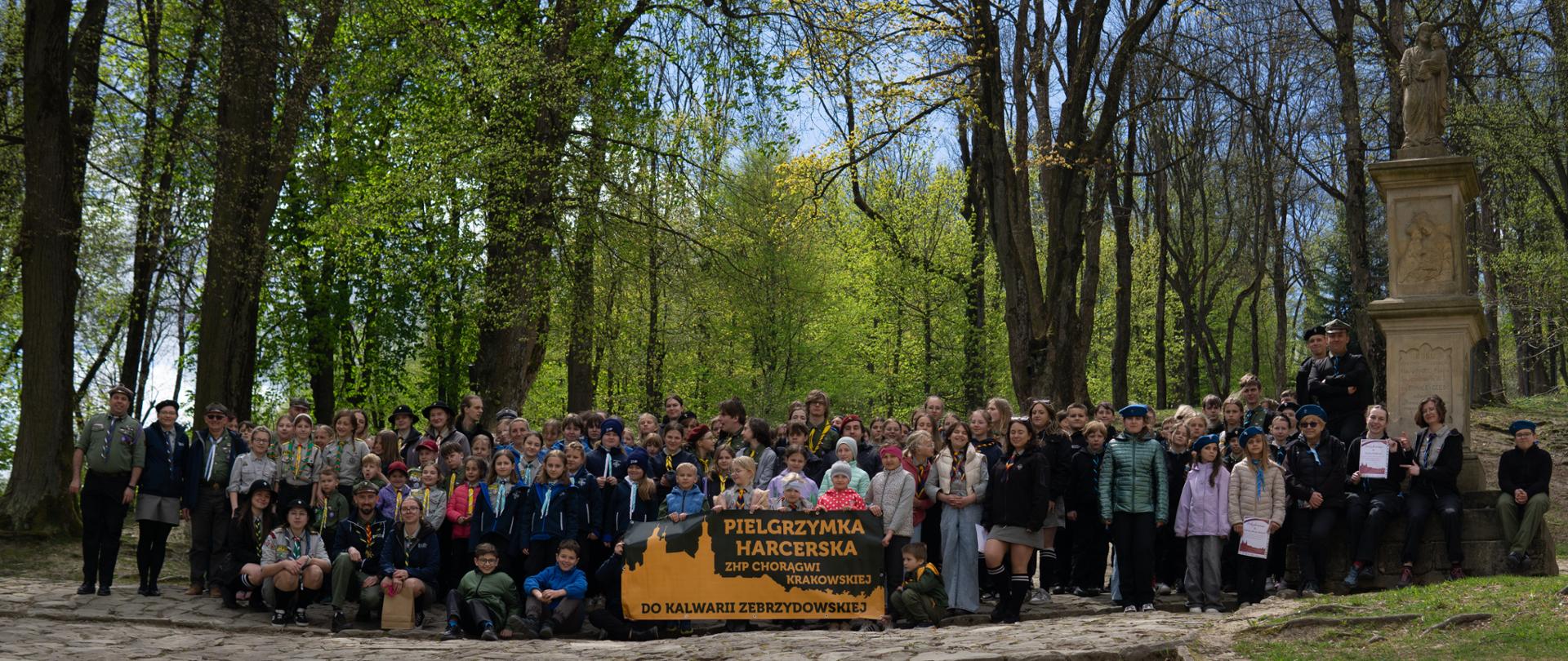 Grupa harcerzy i instruktorów ZHP Chorągwi Krakowskiej zgromadzona na leśnej drodze w Kalwarii Zebrzydowskiej. Uczestnicy w mundurach i chustach stoją wokół banera z napisem „Pielgrzymka Harcerska ZHP Chorągwi Krakowskiej do Kalwarii Zebrzydowskiej”. W tle wiosenna zieleń drzew oraz kamienna figura sakralna. Atmosfera wspólnoty i wydarzenia o charakterze religijnym.