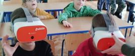 Dzieci siedzą w ławkach w sali lekcyjnej, dwoje z nich ma na oczach interaktywne okulary