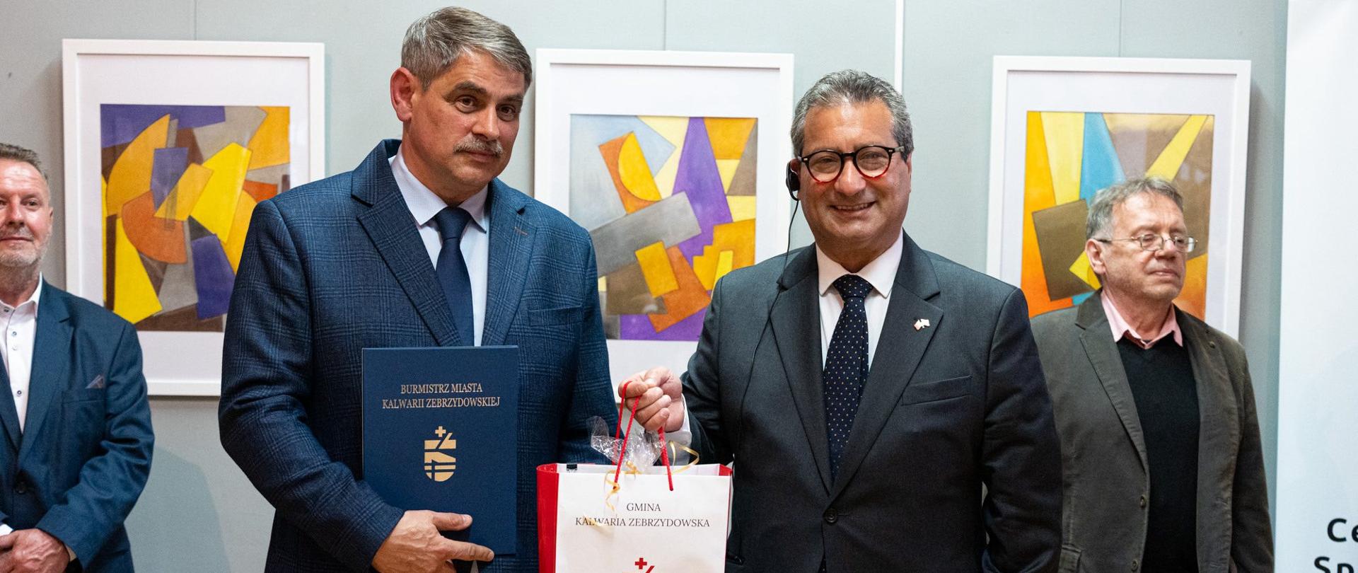 Burmistrz Miasta Kalwarii Zebrzydowskiej Tadeusz Stela wręcza upominek Ambasadorowi Republiki Malty w Polsce dr. Gavinowi Gulii podczas wydarzenia artystycznego w Kalwarii Zebrzydowskiej. Obaj stoją na tle prezentowanych obrazów abstrakcyjnych. W spotkaniu uczestniczą również zaproszeni goście oraz przedstawiciele lokalnych instytucji kultury.