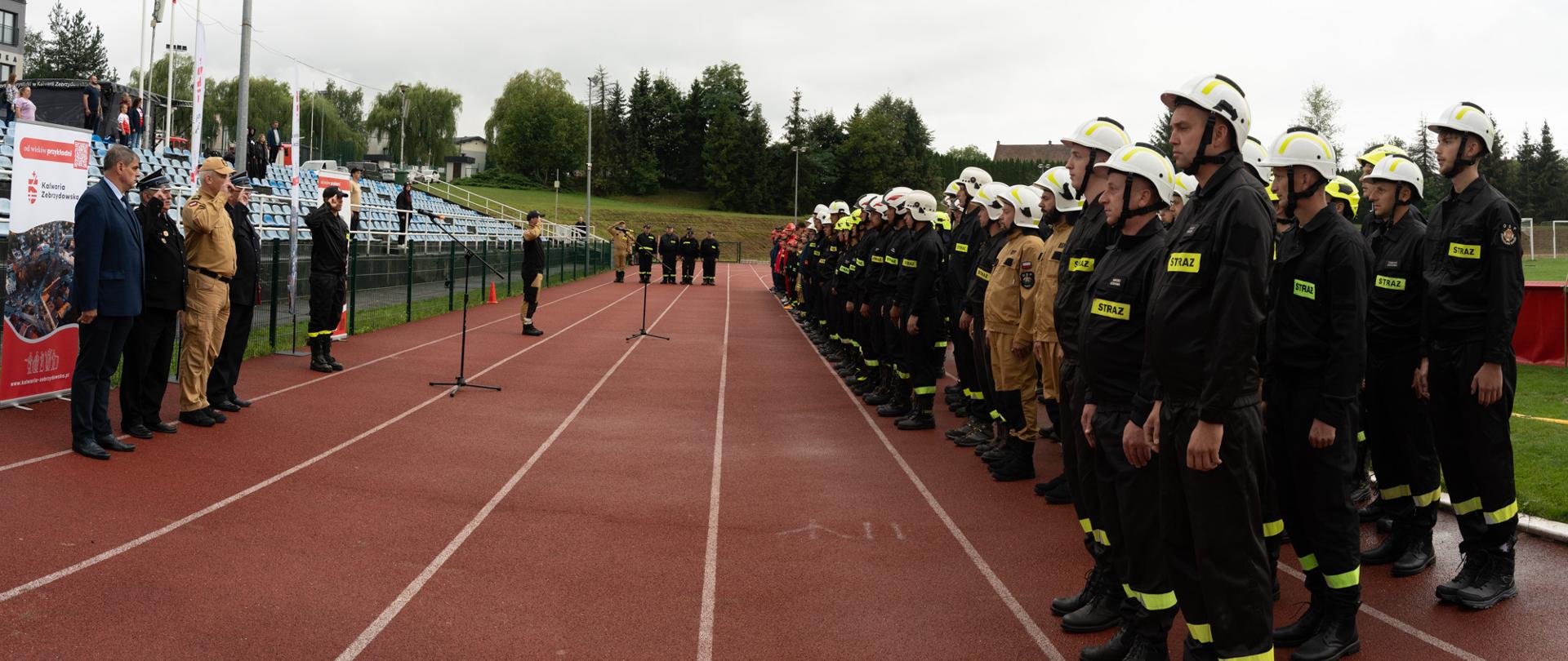 Uczestnicy Gminnych Zawodów Sportowo-Pożarniczych OSP i MDP stoją w szyku na bieżni stadionu MKS Kalwarianka podczas oficjalnej zbiórki. Naprzeciw nich ustawieni są przedstawiciele władz samorządowych i służb pożarniczych. Kalwaria Zebrzydowska, 29 sierpnia 2026 roku. Fot. Andrzej Famielec.