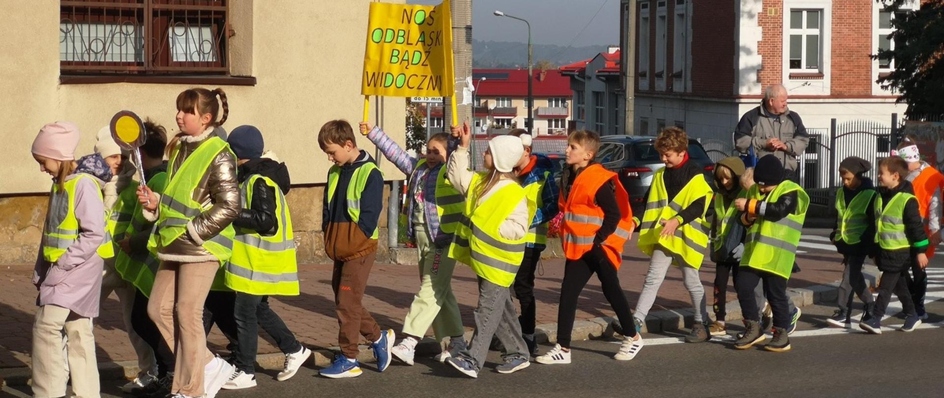 Grupa dzieci ubranych w odblaskowe kamizelki idzie prawą stroną jezdni, niosąc transparent z napisem Noś odblaski bądź widoczny, w tle ściany budynków