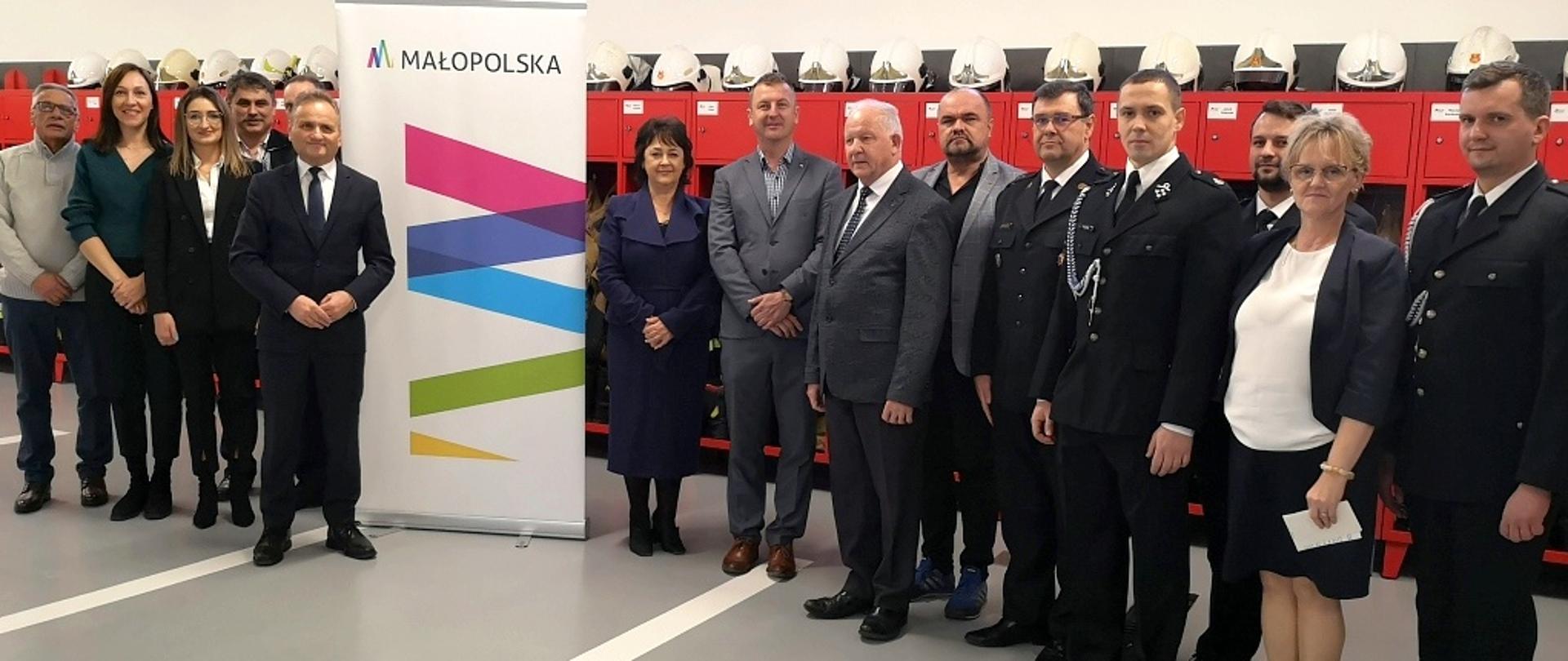 Przedstawiciele samorządu i województwa pozują do zdjęcia w wyremontowanym garażu. Między nimi rollup z logo Województwa Małopolskiego, Z tyłu ściana z dużą ilością okien, pod nią czerwone szafki na odzież strażaków