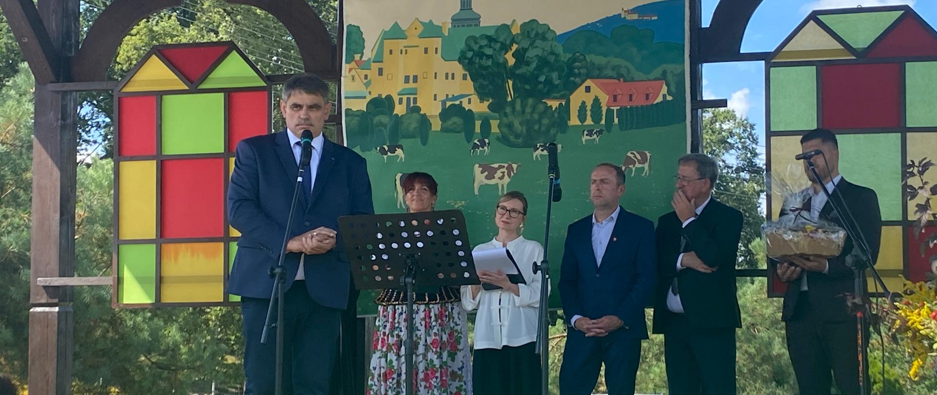 Na plenerowej scenie mężczyzna w granatowym garniturze przemawia do mikrofonu. Za nim stoją uczestnicy uroczystości, a w tle widoczna jest dekoracja przedstawiająca wiejski krajobraz z zabudowaniami i pasącymi się krowami. Scenę zdobią kolorowe elementy oraz dożynkowe kłosy zbóż.
