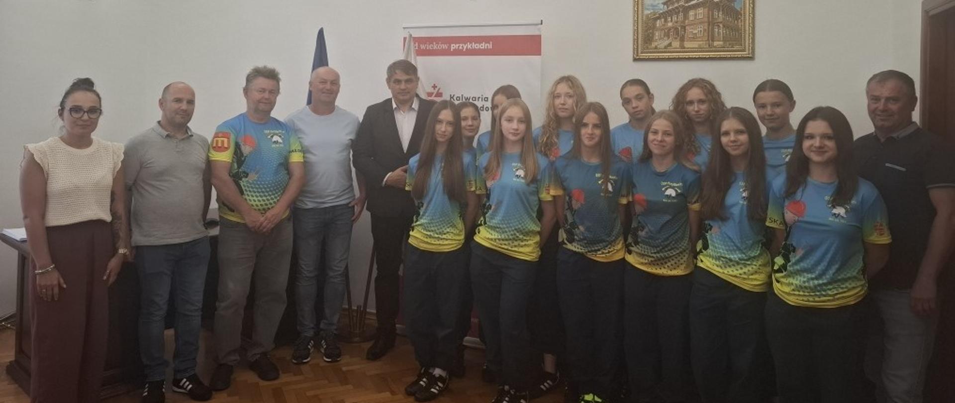 Na zdjęciu widoczna jest grupa osób stojących w pomieszczeniu urzędowym. W tle widać biały baner z herbem oraz napisem „Kalwaria”, na ścianie wisi obraz oraz godło Polski. W pierwszym rzędzie stoi kilkanaście młodych dziewcząt w jednolitych sportowych koszulkach w kolorze niebiesko-żółto-zielonym z nadrukiem balonów. Po bokach i z tyłu znajdują się dorośli – kobieta i kilku mężczyzn, ubrani w stroje codzienne lub eleganckie. Grupa pozuje do wspólnego zdjęcia.