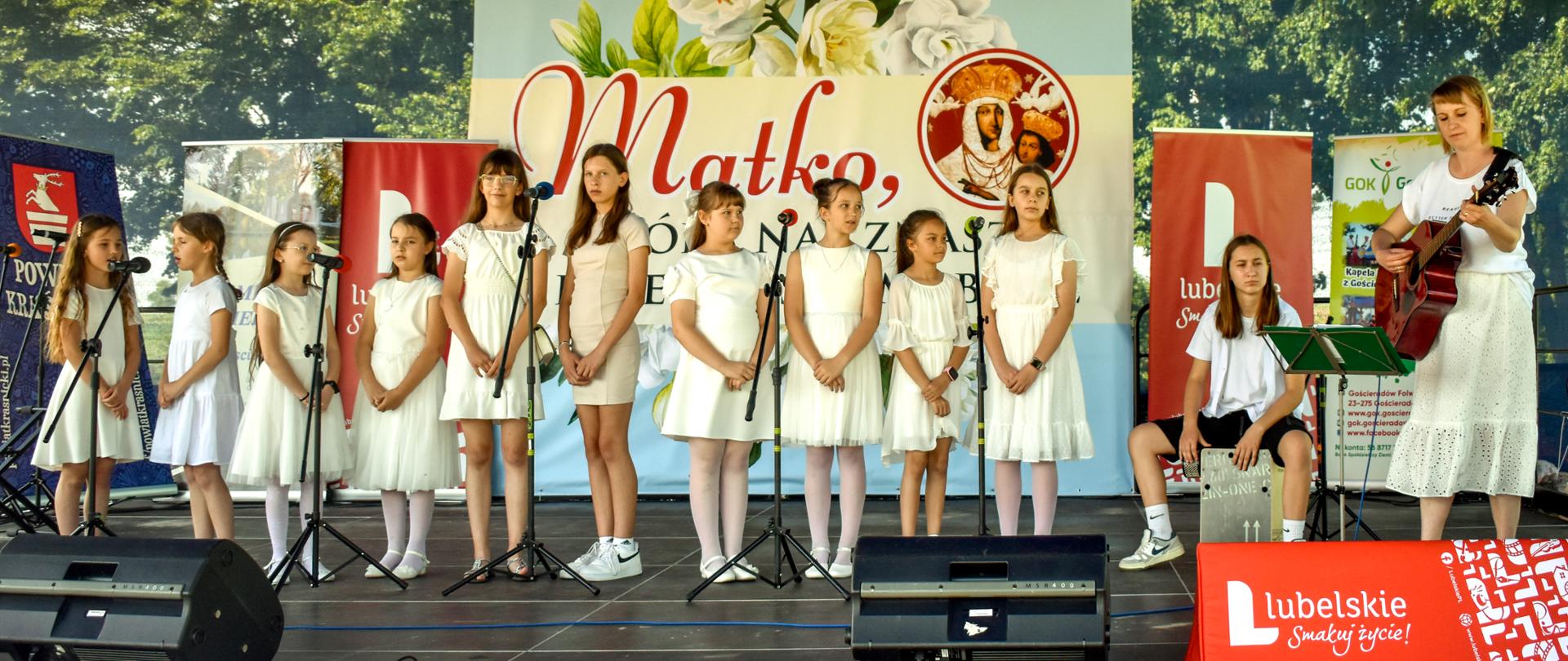 Zdjęcie przedstawia scenę na której stoją śpiewające dziewczynki w jasnych strojach. Jedna z dziewczynek siedzi i gra na instrumencie perkusyjnym. Po prawej stronie stoi kobieta grająca na gitarze. Z tyłu za dziećmi, w centralnej części sceny wisi baner z tytułem festiwalu i grafiką Matki Boskiej Księżomierskiej. Po bokach znajdują się rollupy organizatorów.