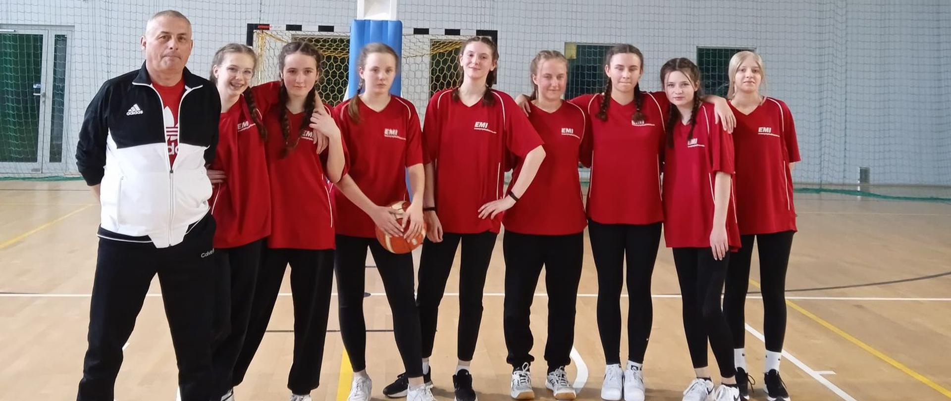 Zdjęcie grupowe koszykarek z trenerem, w tle kosz. 