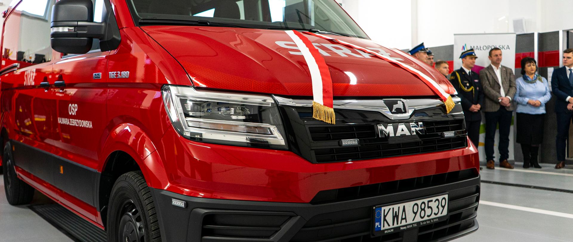 Czerwony przód samochodu straży pożarnej w tle strażak, mężczyzna w jasnym garniturze, kobieta w jasno niebieskiej marynarce, mężczyzna w granatowym garniturze