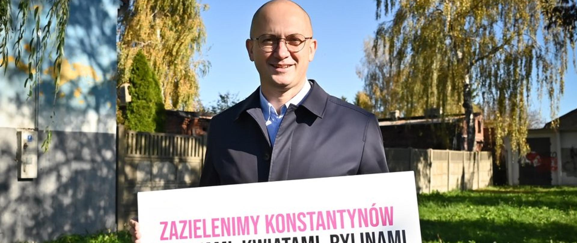 Mężczyzna w okularach stoi na trawie i trzyma tablicę z napisem: ‘Zazielenimy Konstantynów drzewami, kwiatami, bylinami’. W tle zieleń i budynki.