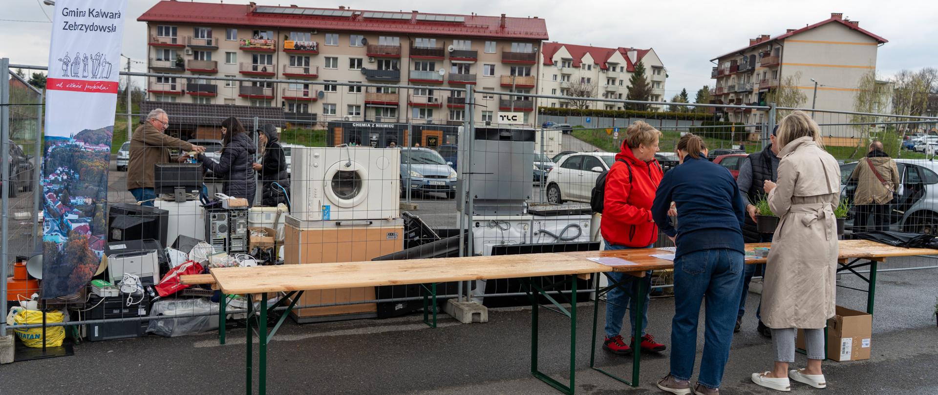 Mieszkańcy oddają elektroodpady podczas gminnej akcji zbiórki – na pierwszym planie stoły, przy których uczestnicy odbierają sadzonki roślin, w tle widoczne zużyte sprzęty AGD i RTV (m.in. pralka, komputery), ogrodzony teren oraz zabudowa mieszkaniowa Kalwarii Zebrzydowskiej.