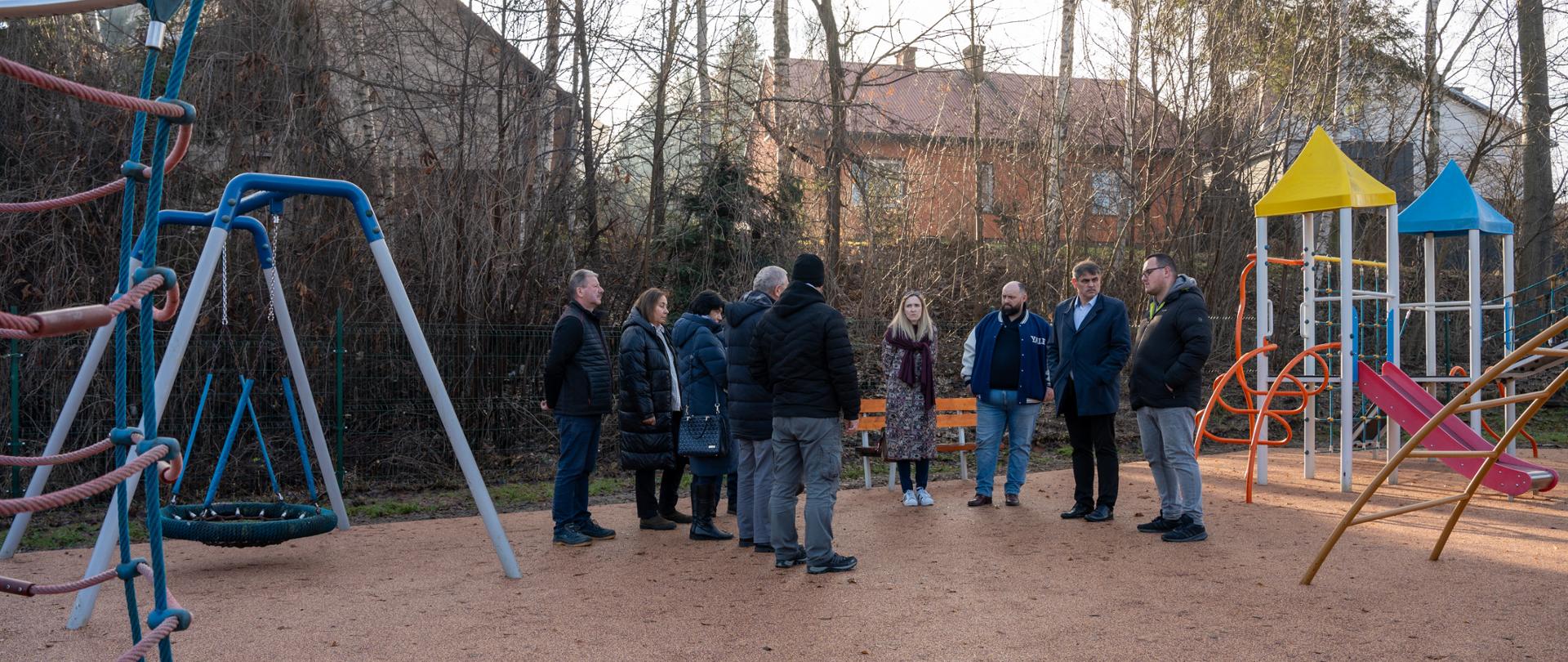 Grupa przedstawicieli samorządu i zaproszonych osób stoi na nowo zmodernizowanym placu zabaw, prowadząc rozmowę podczas odbioru inwestycji. W tle widoczne są kolorowe urządzenia zabawowe, huśtawki oraz zestaw ze zjeżdżalniami, a całość znajduje się na terenie otoczonym drzewami i zabudową jednorodzinną.