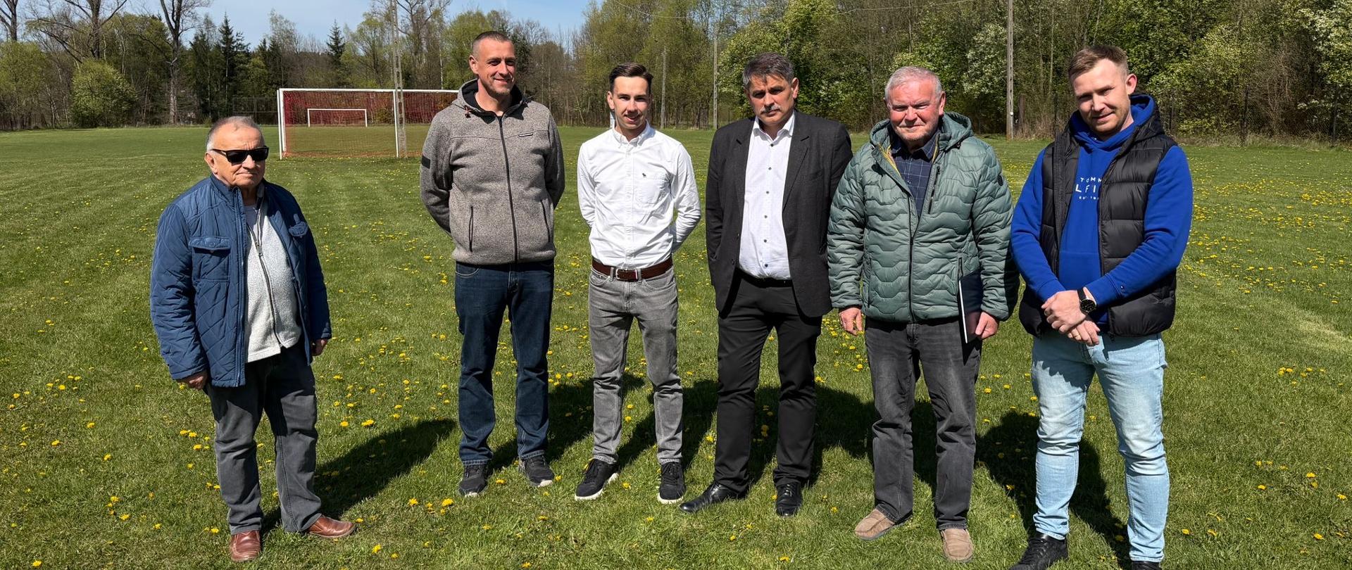 Siedmiu mężczyzn stoi na trawiastym boisku sportowym w słoneczny dzień. W tle widoczna jest bramka piłkarska, słupy oświetleniowe oraz drzewa z wiosenną zielenią. Mężczyźni ubrani są w casualowe i półformalne stroje – kurtki, bluzy i koszule – i stoją obok siebie, patrząc w stronę aparatu. Na murawie widoczne są żółte kwiaty mniszka lekarskiego. Atmosfera zdjęcia sugeruje spotkanie w terenie związane z infrastrukturą sportową lub lokalną inwestycją.