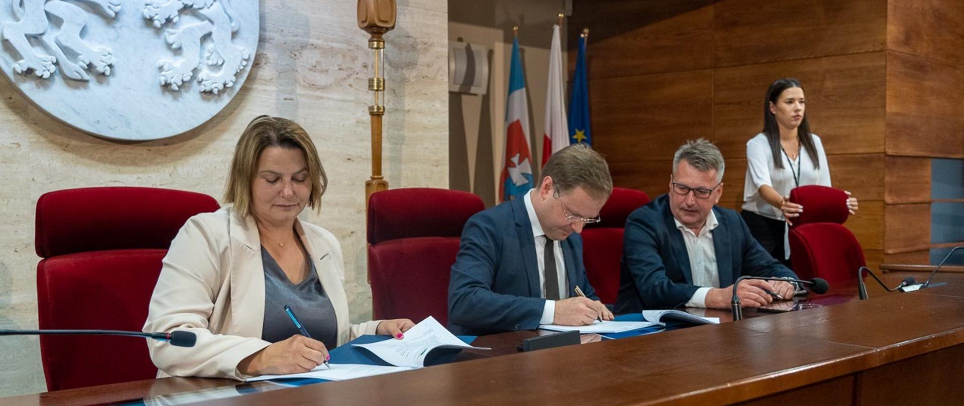Na fotografii znajduję się dwóch mężczyzn oraz dwie kobiety. Od lewej strony kobieta ubrana w szarą koszulkę oraz biały żakiet. Obok niej dwóch mężczyzn ubranych w granatowe garnitury i białe koszule. Mężczyźni posiadają okulary. Za nimi stoi kobieta w białej bluzce, obejmująca dłońmi czerwony fotel. Osoby podpisują umowy na dużym drewnianym biurku. Siedzą na dużych czerwonych fotelach oraz każdy z nich w dłoniach posiada pióro. Za nimi na jasno marmurowej ścianie znajduje się biały wykuty herb Urzędu Marszałkowskiego. Obok stoją flagi.
Autor zdjęć Anna Magda - Biuro Prasowe UMWP
