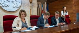 Na fotografii znajduję się dwóch mężczyzn oraz dwie kobiety. Od lewej strony kobieta ubrana w szarą koszulkę oraz biały żakiet. Obok niej dwóch mężczyzn ubranych w granatowe garnitury i białe koszule. Mężczyźni posiadają okulary. Za nimi stoi kobieta w białej bluzce, obejmująca dłońmi czerwony fotel. Osoby podpisują umowy na dużym drewnianym biurku. Siedzą na dużych czerwonych fotelach oraz każdy z nich w dłoniach posiada pióro. Za nimi na jasno marmurowej ścianie znajduje się biały wykuty herb Urzędu Marszałkowskiego. Obok stoją flagi.
Autor zdjęć Anna Magda - Biuro Prasowe UMWP