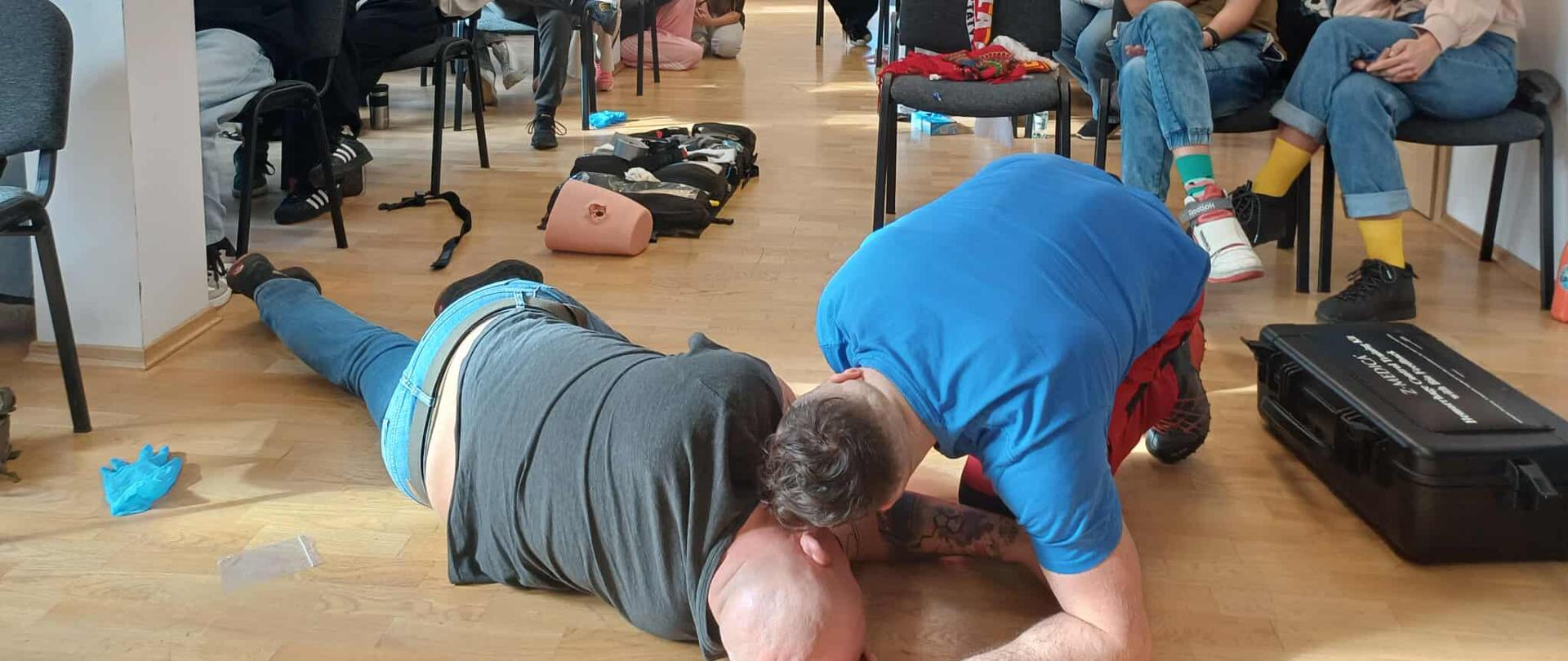 W jasnej sali szkoleniowej trwa pokaz udzielania pierwszej pomocy. Ratownik w niebieskiej koszulce prezentuje techniki ratunkowe na osobie leżącej na podłodze, podczas gdy kilkanaście osób siedzących na krzesłach i stojących w tle uważnie obserwuje przebieg szkolenia. Na podłodze widoczne są elementy wyposażenia medycznego oraz fantom do ćwiczeń. Atmosfera skupienia wskazuje na praktyczne zajęcia z zakresu ratownictwa i pierwszej pomocy.