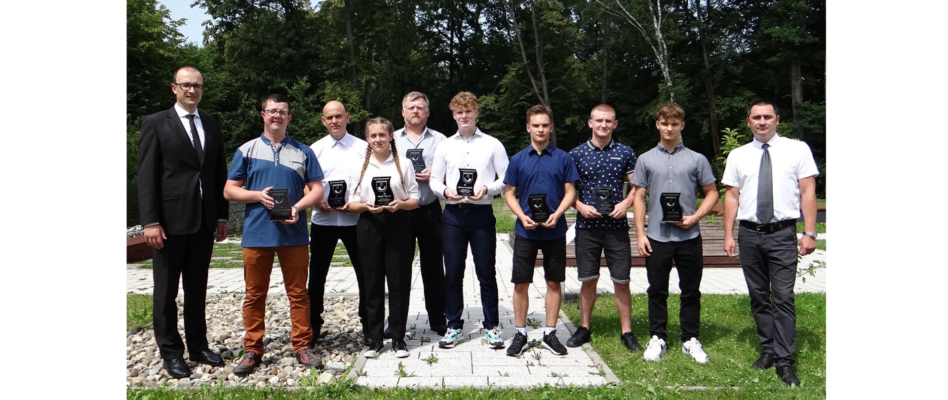 Laureaci nagród w towarzystwie Wójta Grzegorza Sikorskiego i Przewodniczącego Rady Gminy Sławomira Kolondry. Osoby stoją w plenerze. Wokół trawa, z tyłu drzewa liściaste