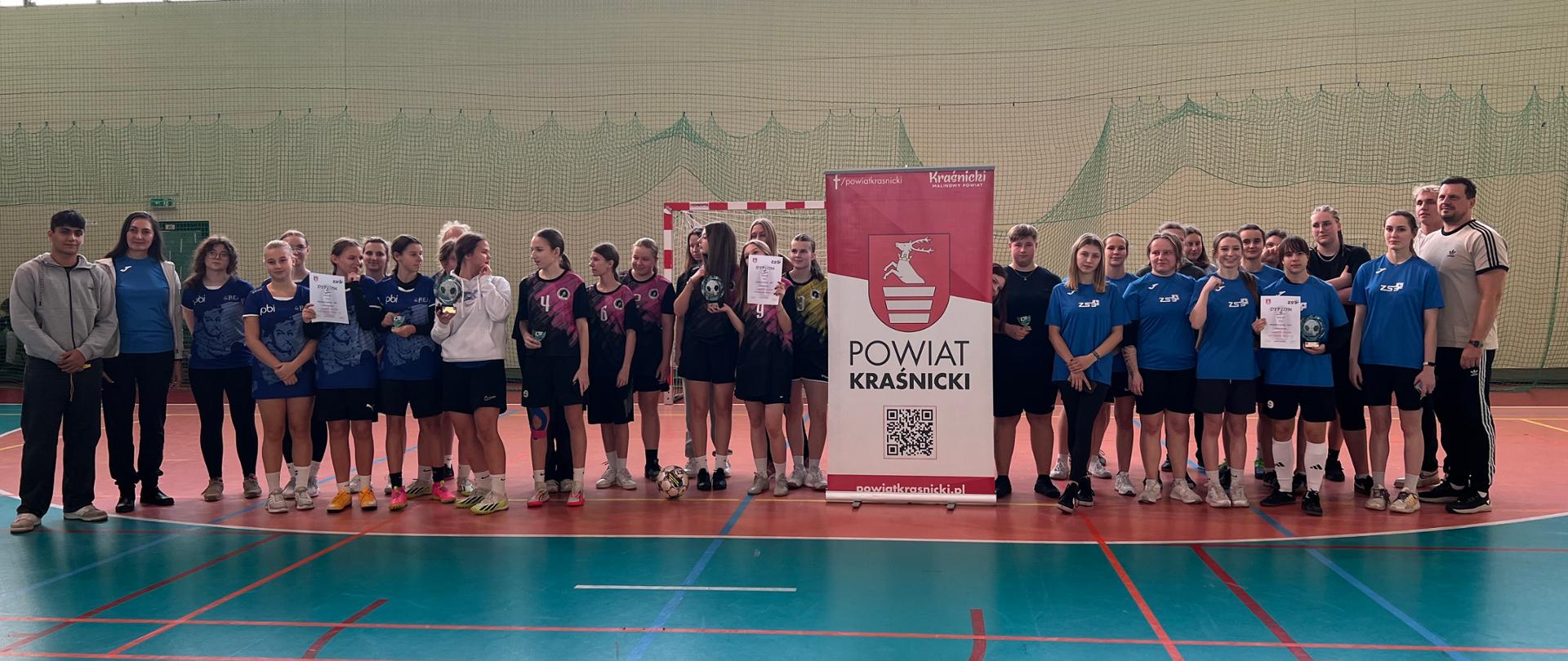 Uczestniczki turnieju stojące wraz z opiekunami ze szkół i organizatorem turnieju na hali sportowej. Pośrodku nich ustawiony jest rollup Starostwa Powiatowego w Kraśniku