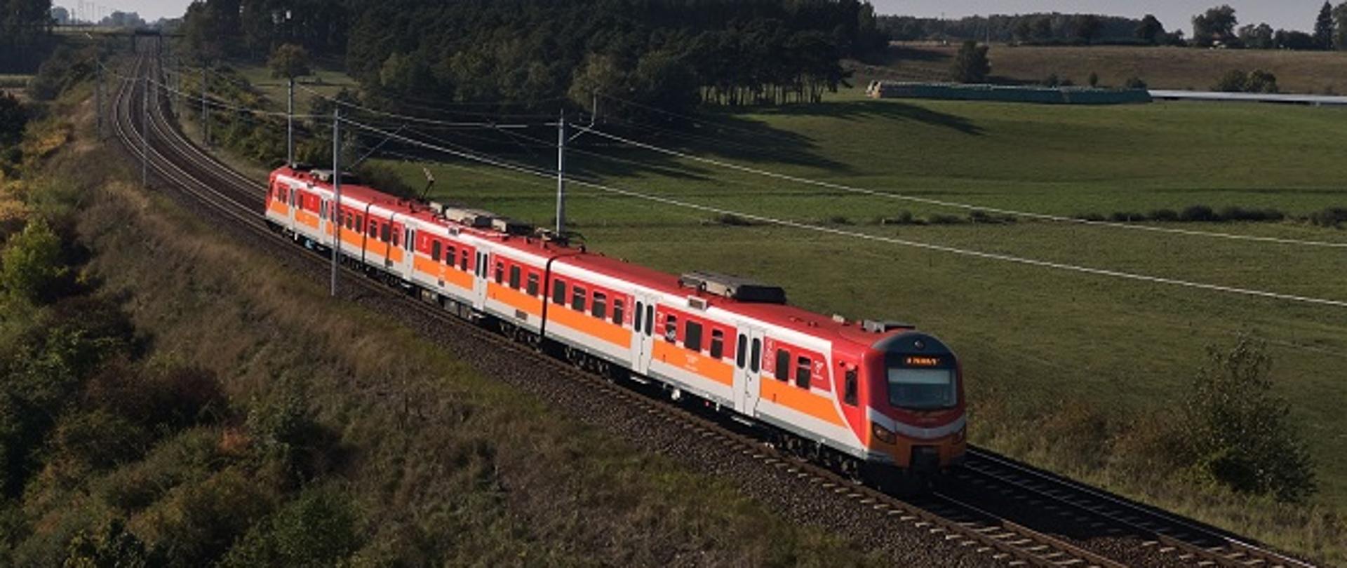 Czerwono-biały pociąg pasażerski jadący po torach kolejowych przez zielone pola i lekko pagórkowaty krajobraz wiejski. Zdjęcie wykonane z lotu ptaka w słoneczny dzień, w tle widoczny las oraz otwarta przestrzeń.
