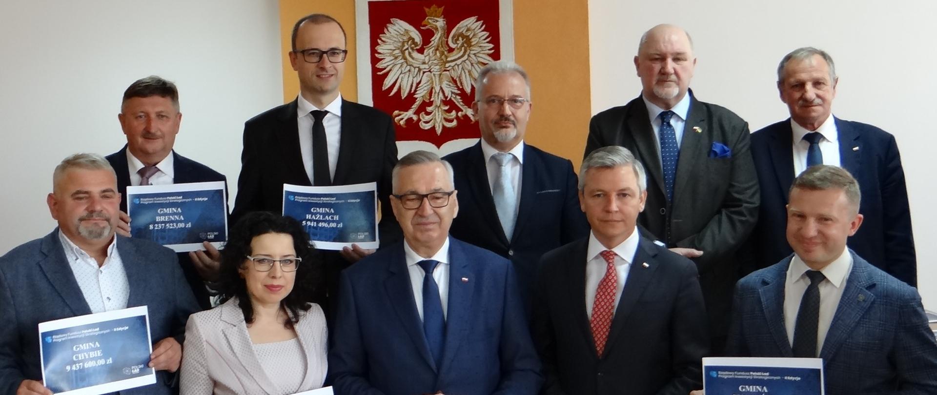 Grupa biorących udział w spotkaniu samorządowców z terenu Powiatu Cieszyńskiego z promesami Polskiego Ładu. W ostatnim rzędzie drugi od lewej Wójt Gminy Hażlach Grzegorz Sikorski. Osoby stoją w dwóch rzędach. Z tyłu polskie godło 