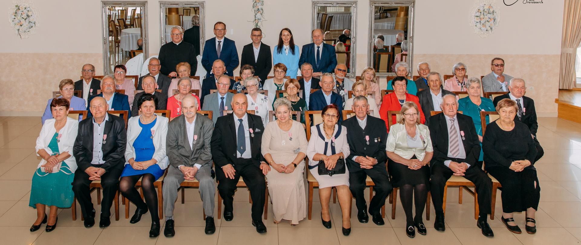 Zdjęcie przedstawia uczestników Jubileusz 50-lecia Pożycia Małżeńskiego, siedzących na krzesłach oraz stojących za nimi Ks.Dariusza Ostrowskiego, urzędników Urzędu Stanu Cywilnego wraz z Wójtem Gminy Andrzejem Dziwiszem i posłem na Sejm RP Edwardem Siarką pozujących do zdjęcia na tle ściany z lustrami w restauracji.