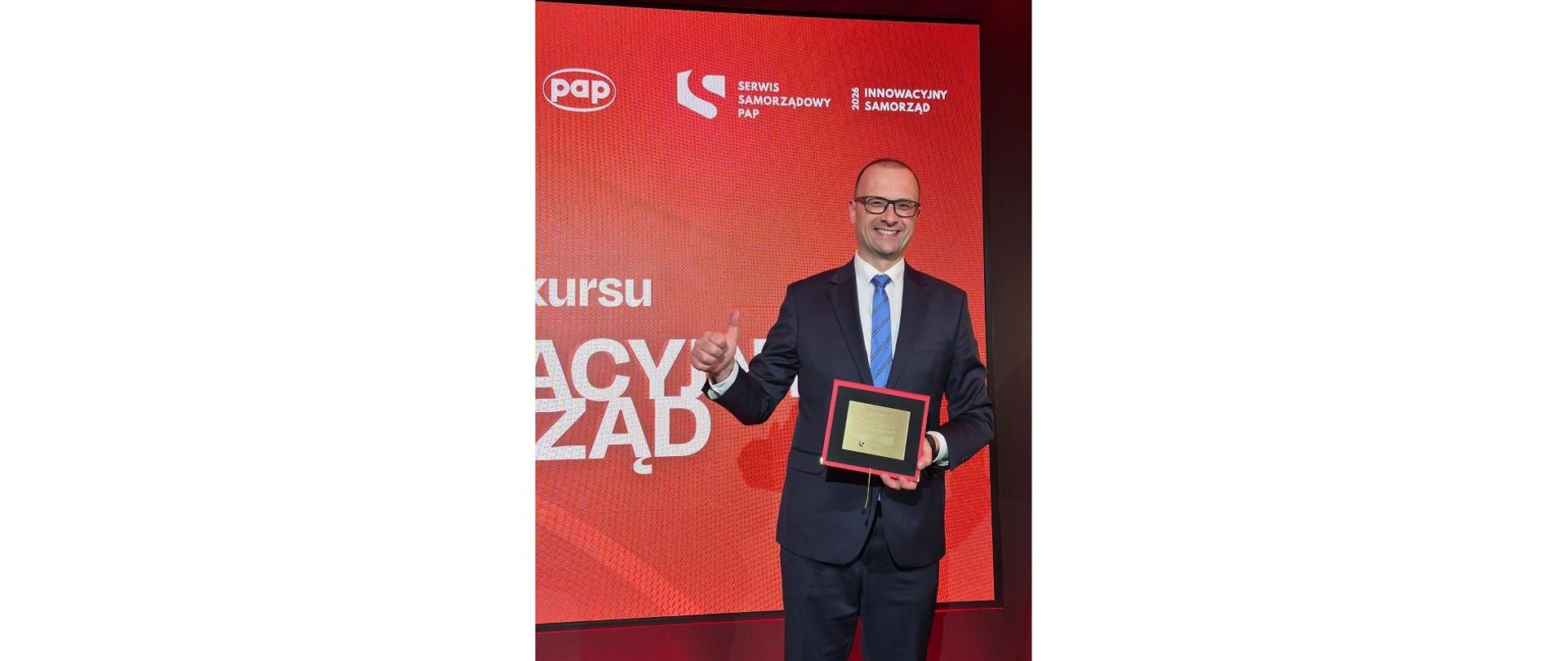 Wójt Grzegorz Sikorski odbiera w Warszawie wyróżnienie przyznane w konkursie Serwisu Samorządowego Polskiej Agencji Prasowej. Uśmiechnięty szeroko mężczyzna z podniesionym do góry kciukiem prezentuje otrzymane wyróżnienie