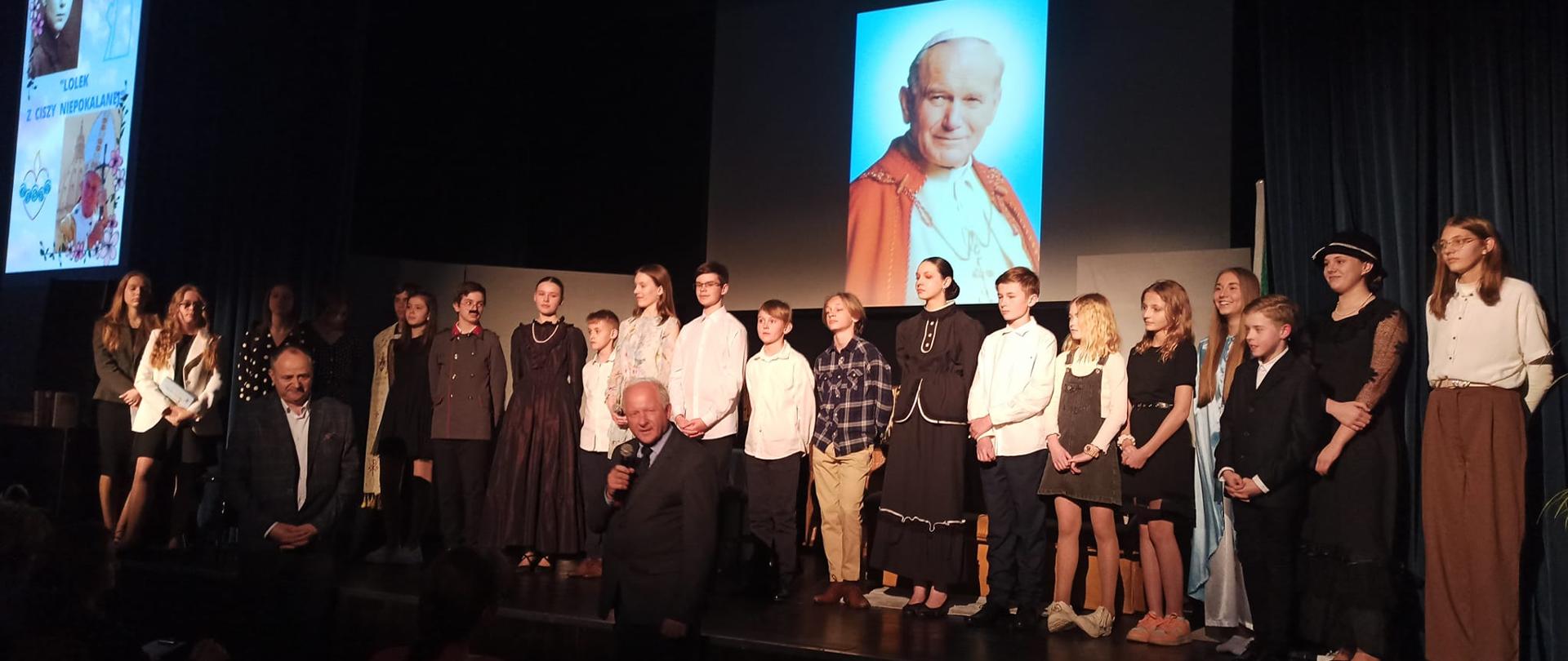 Na oświetlonej scenie w rzędzie stoją młodzi aktorzy teatralni. Za nimi na ekranie portret Jana Pawła II. Przed sceną burmistrz wypowiadający się do mikrofonu.