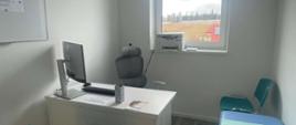Na pierwszym planie znajduje się niebieska kozetka, białe biurko oraz szare krzesło biurowe w tle sprzęt elektroniczny oraz w tle okno