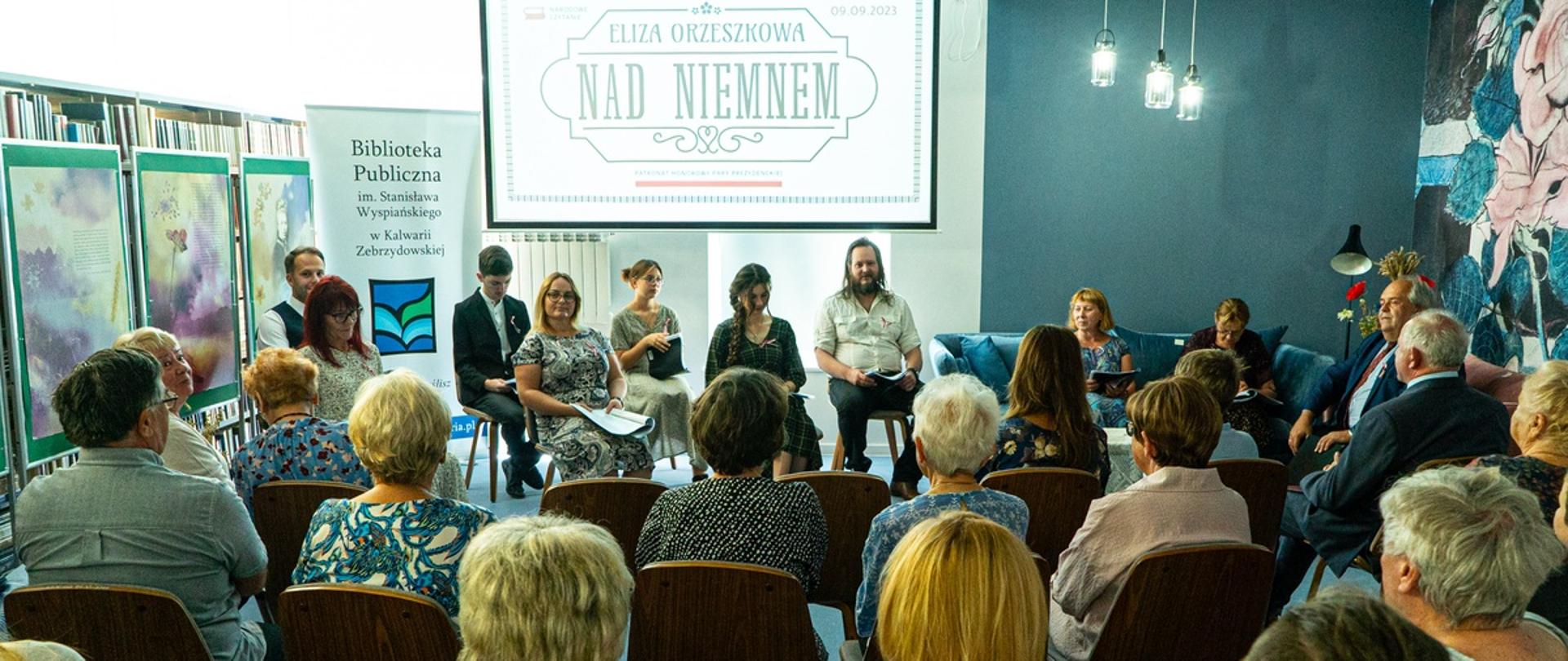 W pomieszczeniu biblioteki na pierwszym planie siedzą tyłem do aparatu słuchacze Narodowego Czytania, przed nimi osoby czytające teksty. W tle widoczne grafiki okolicznościowe i ekran z tematyką tegorocznego czytania.