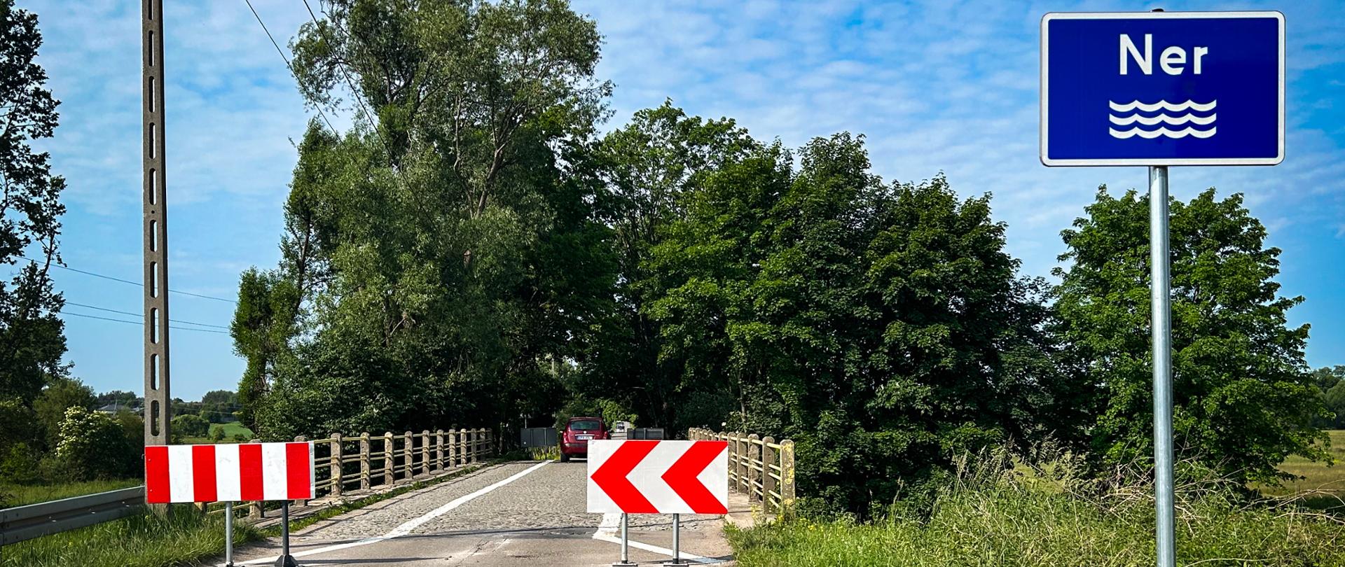 Zamknięty wjazd na most przez rzekę Ner. Na jezdni ustawione są biało-czerwone bariery i znaki kierunkowe. Po prawej stronie widoczny niebieski znak z nazwą rzeki 'Ner' i symbolem fal. W tle drzewa, zieleń i niebo z lekkim zachmurzeniem.