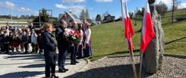 Delegacja z kwiatami przed pomnikiem, kobieta i dwóch mężczyzn. Obok stoją poczty sztandarowe i biało-czerwone flagi. W tle grupa dzieci.