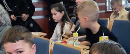 Uczestnicy turnieju w oczekiwaniu na wyniki. Na pierwszym planie dwójka chłopców w strojach strażackich, w tle widać inne dzieci które również uczestniczyły w turnieju. Wszyscy siedzą przy stolikach korzystając z przerwy piją soczki oraz jedzą batonika od organizatorów.