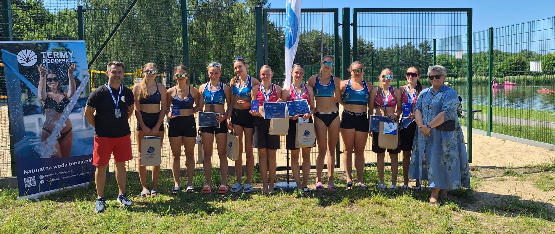 Grupowe zdjęcie uczestniczek turnieju siatkówki plażowej
Grupa młodych zawodniczek w strojach sportowych pozuje z medalami, dyplomami i nagrodami na tle boiska do siatkówki plażowej. Po bokach stoją organizatorzy – mężczyzna w czarnym podkoszulku i kobieta w niebieskiej sukience. W tle widoczna jest siatka, drzewa oraz zbiornik wodny z różowymi rowerami wodnymi.