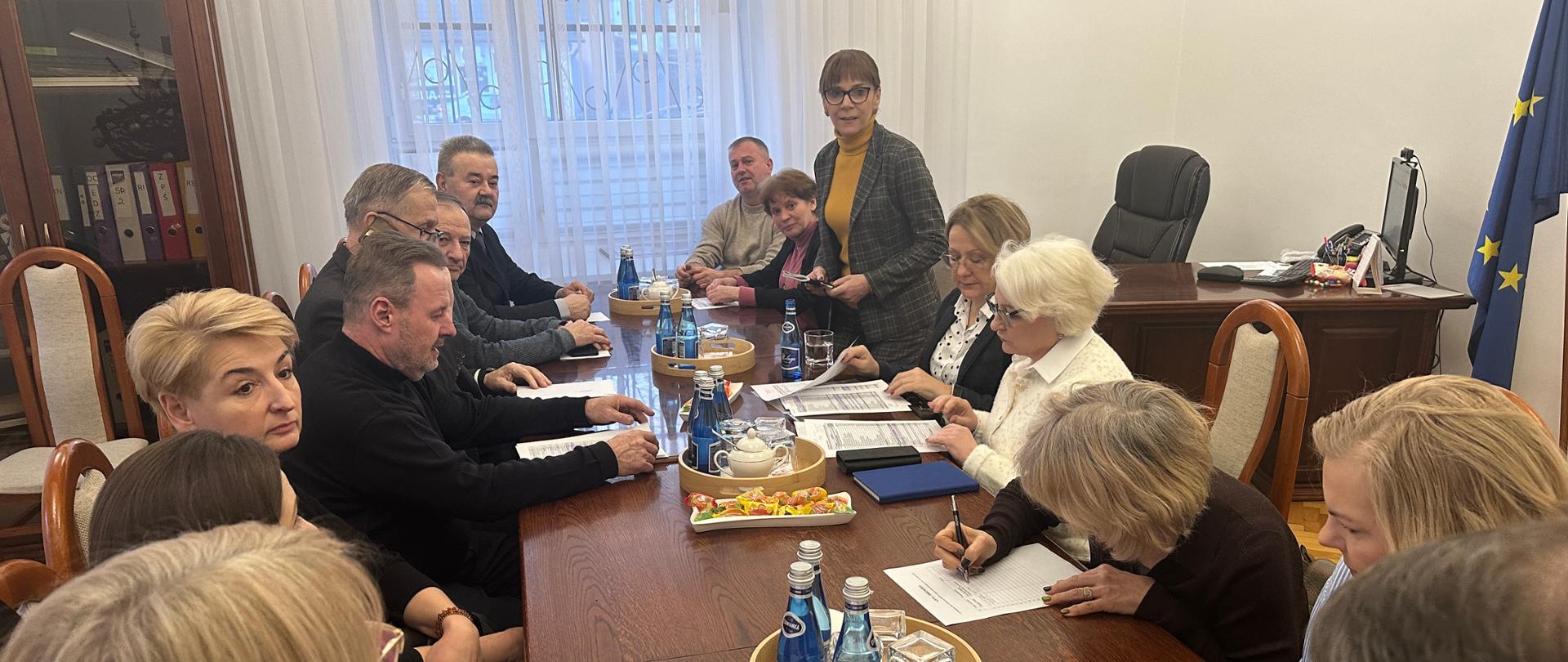 Grupa kilkunastu osób zasiada przy długim, drewnianym stole w sali urzędowej podczas oficjalnego posiedzenia. Uczestnicy przeglądają dokumenty, podpisują listy i prowadzą rozmowy. Na stole znajdują się teczki z papierami, długopisy, notatniki, woda mineralna i drobny poczęstunek. W tle widoczne są regały z aktami, biurko oraz flaga Unii Europejskiej, co podkreśla formalny charakter spotkania.