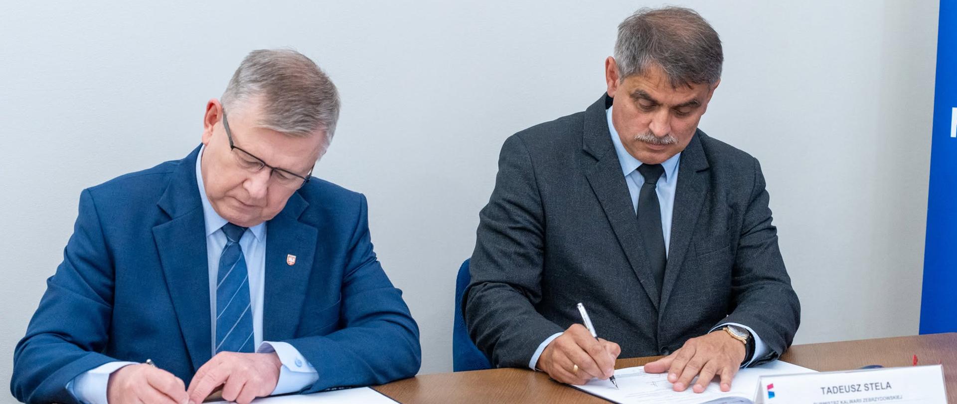 Dwaj mężczyźni w garniturach (marszałek i burmistrz) siedzą przy stole i podpisują dokumenty
