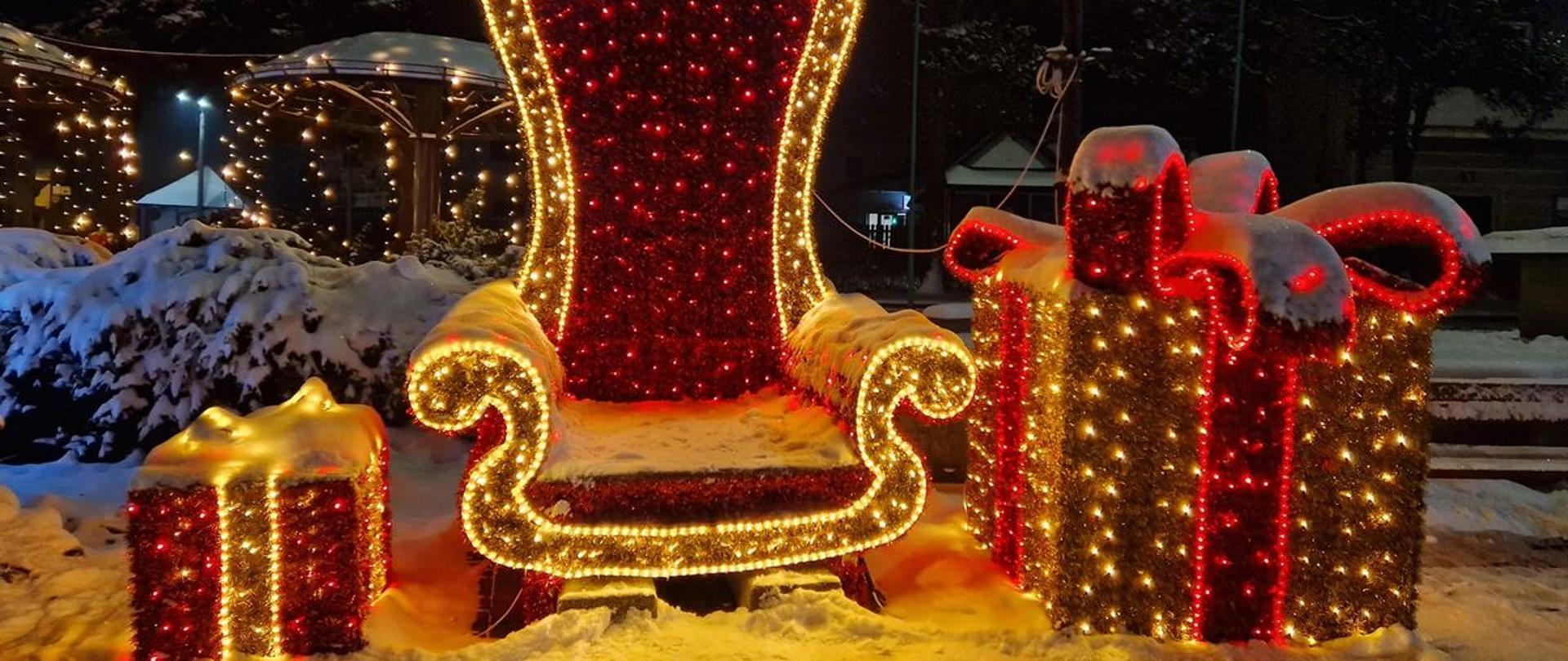Miejska dekoracja świąteczna w postaci dwóch prezentów i fotela dla Mikołaja, mieniących się po zmroku żółtymi i czerwonymi światełkami.