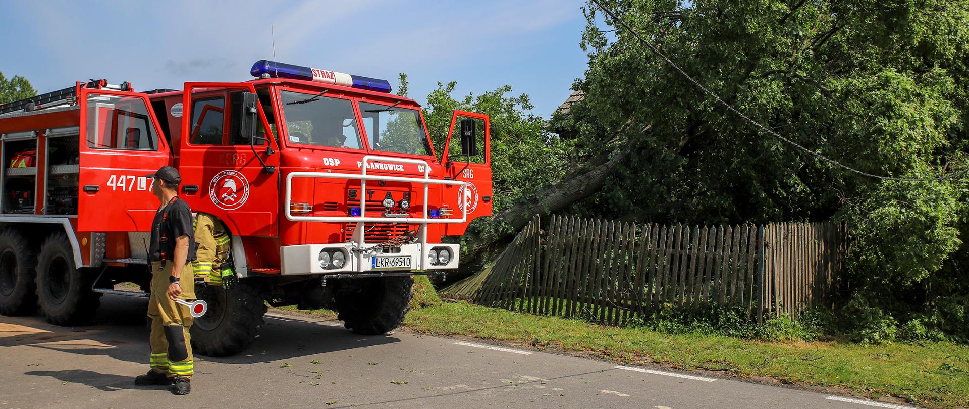 Na zdjęciu wóz strażacki oraz jego załoga zabezpieczają teren wokół domu, na który upadło powalone przez burzę drzewo.