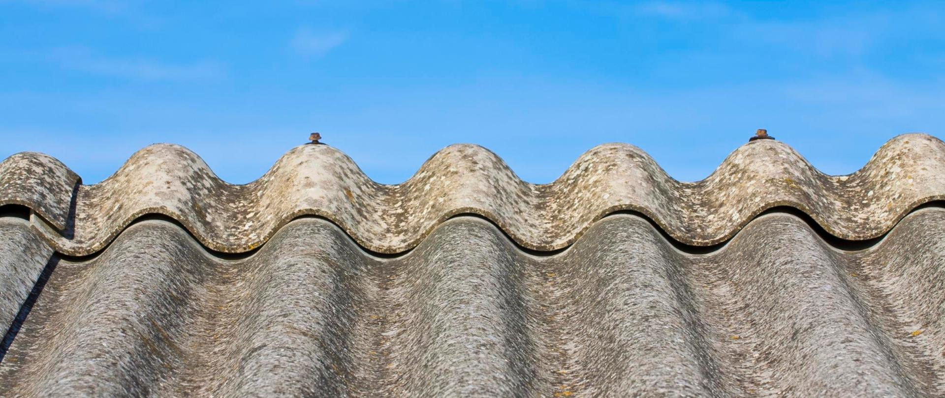 Zbliżenie na falisty dach z szarych płyt azbestowo-cementowych (eternit) na tle błękitnego nieba.