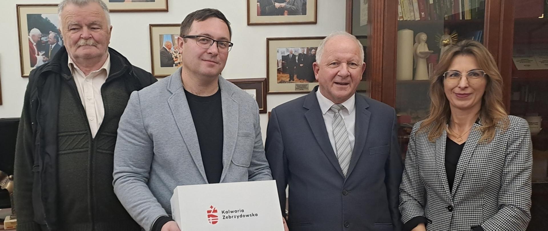 Trzech mężczyzn stoi, od lewej strony najwyższy mężczyzna z wąsami w rozpiętej kurtce, swetrze i jasnej koszuli, kolejny mężczyzna w okularach, rozpiętej jasnej marynarce i czarnym podkoszulku prezentuje trzymając przed sobą pudełko białe z logo Kalwarii Zebrzydowskiej oraz napisem Kalwaria Zebrzydowska, kolejny mężczyzna, burmistrz, z siwymi włosami, uśmiechnięty w dopasowanej grafitowej marynarce, jasnej koszuli i krawatem w poprzeczne paski, najbardziej z prawej stoi kobieta z kasztanowymi włosami po ramiona, w okularach w marynarce w drobną kratę biało czarną oraz czarną bluzeczką