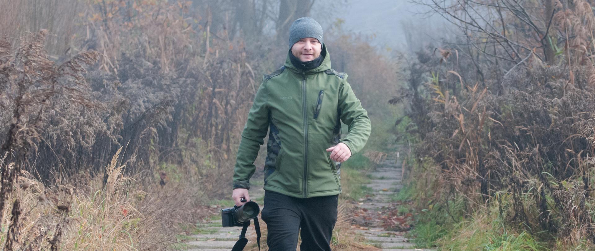 Mężczyzna w zielonej kurtce, czapce i kaloszach idzie ścieżką wśród jesienno-zimowej roślinności, trzymając w ręce aparat fotograficzny z teleobiektywem.