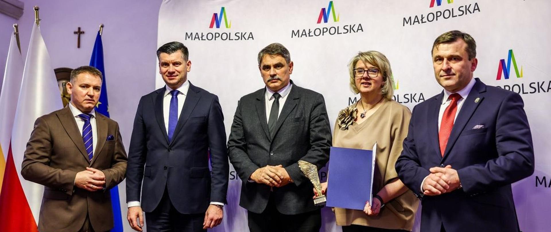 Na zdjęciu stoi pięć osób w eleganckich strojach, ustawionych na tle ścianki z logo „Małopolska”. W środku znajduje się Burmistrz Kalwarii Zebrzydowskiej Tadeusz Stela, trzymający statuetkę. Obok niego stoi kobieta z teczką i dyplomem oraz trzech mężczyzn w garniturach, w tym przedstawiciele władz województwa. Po lewej stronie widoczne są flagi Polski i Unii Europejskiej. Zdjęcie ma charakter oficjalny i dokumentuje wręczenie wyróżnień podczas uroczystości samorządowej.