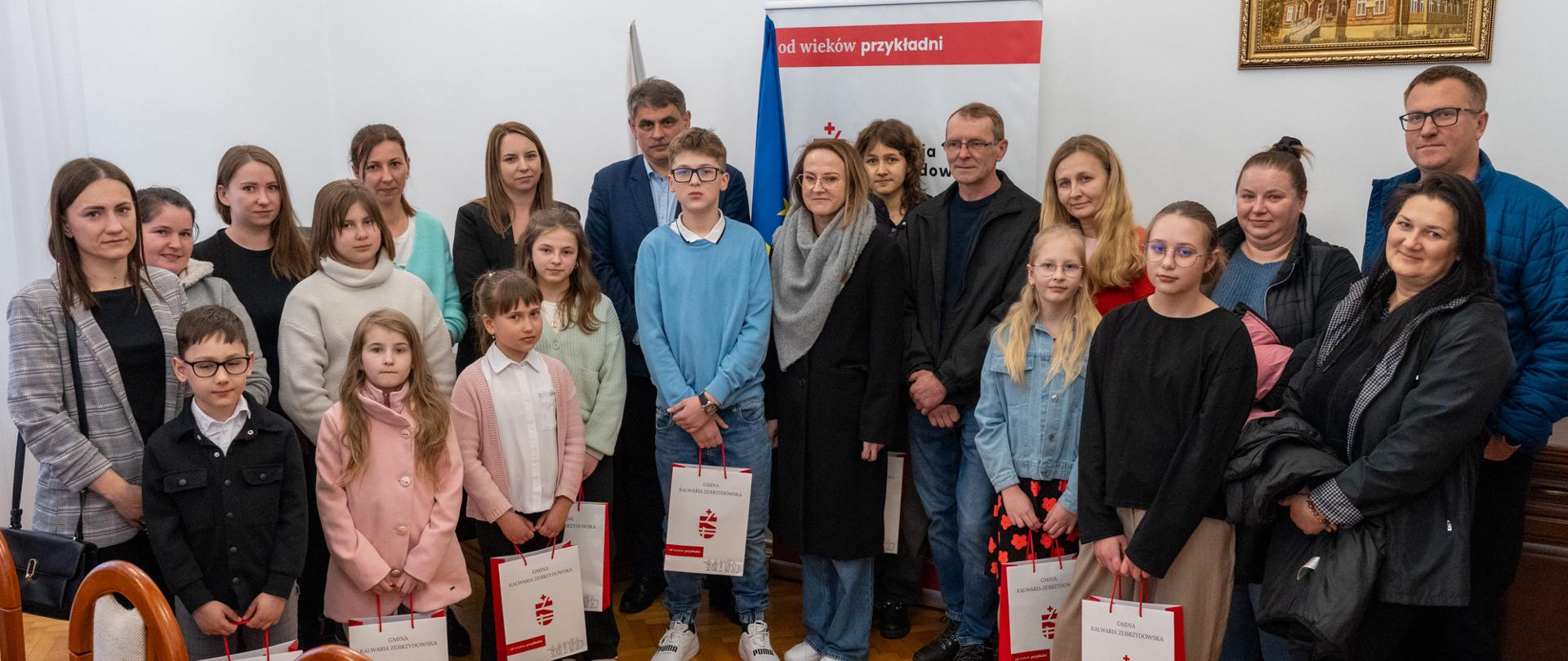 Grupa dzieci i dorosłych stoi w sali urzędu podczas wręczenia nagród. Uczniowie trzymają torby z upominkami z logo Gminy Kalwaria Zebrzydowska. W tle widoczna flaga Polski i Unii Europejskiej oraz roll-up z napisem „od wieków przykładni”. Uczestnicy ustawieni są do wspólnego zdjęcia, panuje uroczysta, ale serdeczna atmosfera.