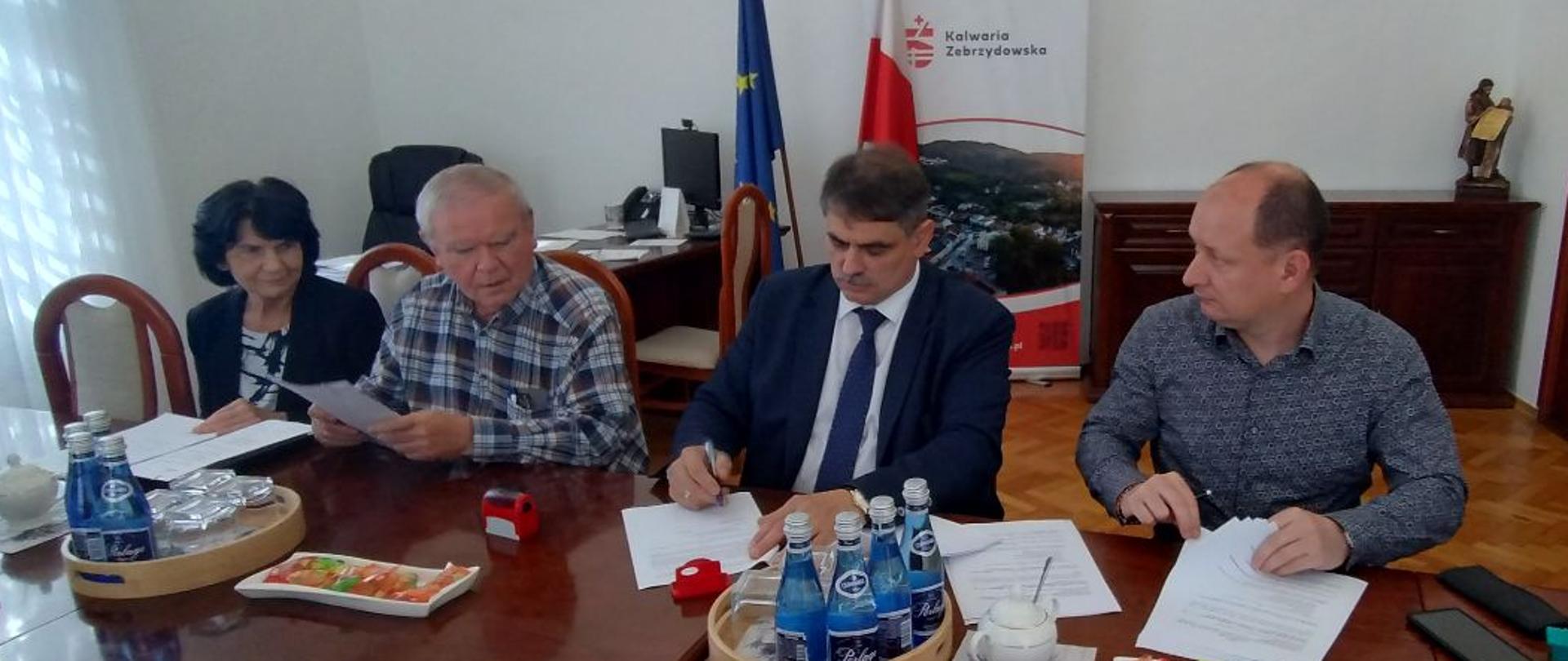 Przy stole na którym stoją butelki wody mineralnej pięć osób podpisuje dokumenty, od lewej strony siedzą kobieta mająca czarne włosy i trzymająca plik papierów, następnie mężczyzna o siwych włosach w koszuli w kratę trzyma w rękach plik kartek, następnie mężczyzna w garniturze i ciemnym krawacie, brunet podpisuje dokumenty, dalej mężczyzna z widoczna łysiną trzyma plik kartek, następnie w prawym rogu siedzi mężczyzna brunet z wąsami, który przygląda się całej sytuacji. 