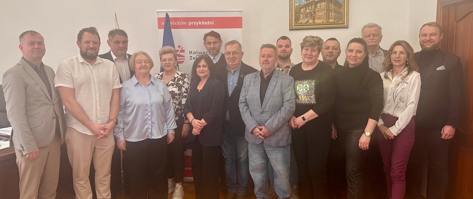 Grupa kilkunastu osób – kobiet i mężczyzn – stoi do wspólnego zdjęcia w gabinecie urzędowym. W tle widoczny jest godło Polski na ścianie, flaga oraz roll-up z herbem i nazwą Kalwarii Zebrzydowskiej. Uczestnicy ustawieni są w dwóch rzędach, ubrani w stroje półformalne i eleganckie. Pomieszczenie ma jasne ściany, drewnianą podłogę oraz obraz przedstawiający zabytkowy budynek.