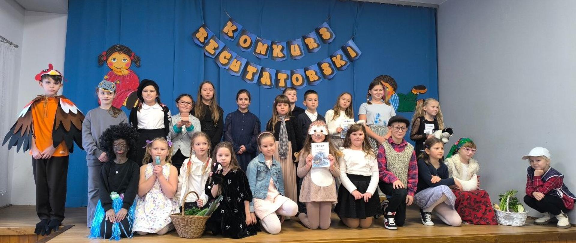 Grupa około trzydziestu dzieci stoi i siedzi na scenie szkolnej podczas konkursu recytatorskiego. Uczniowie mają różnorodne stroje — zarówno codzienne, jak i kostiumy sceniczne — a za nimi znajduje się niebieskie tło z napisem Konkurs Recytatorski wykonanym z dekoracyjnych liter. Dwoje dużych rysunkowych bohaterów zdobi boki sceny. Dzieci pozują wspólnie do zdjęcia grupowego.
