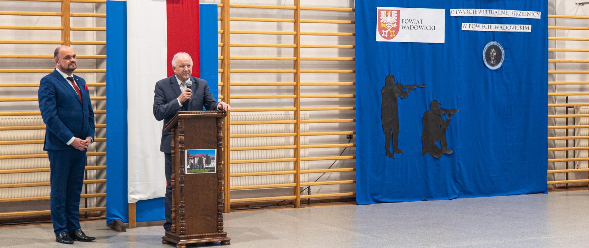 W sali gimnastycznej po lewej stronie mężczyzna w granatowym garniturze obok przy mównicy starszy pan przemawia z mównicy, po prawej stronie gazetka z herbem powiatu wadowickiego oraz żołnierzy. 