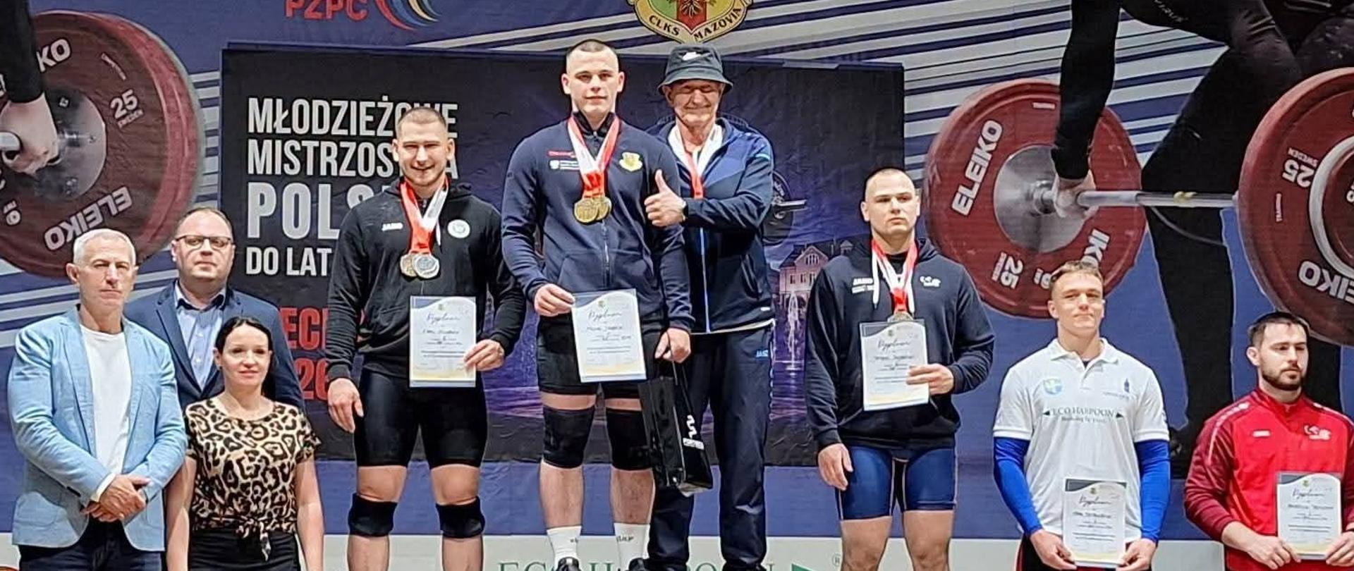 Zawodnicy stoją na podium podczas ceremonii dekoracji na Młodzieżowych Mistrzostwach Polski w podnoszeniu ciężarów. W tle widoczny baner zawodów, a przed podium sztanga z obciążeniami.